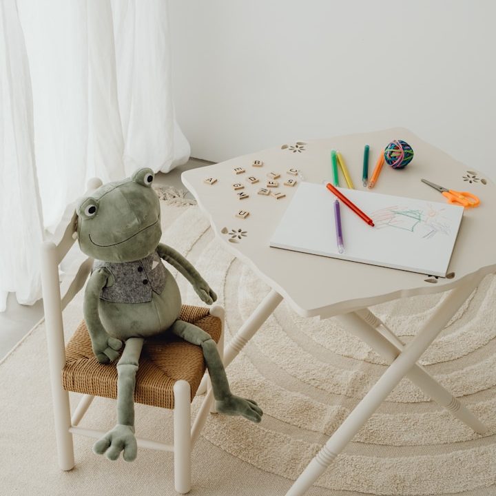 Therapie für Kinder Göppingen Ein grüner Plüschfrosch sitzt auf einem Stuhl neben einem Tisch mit Bastelmaterialien.