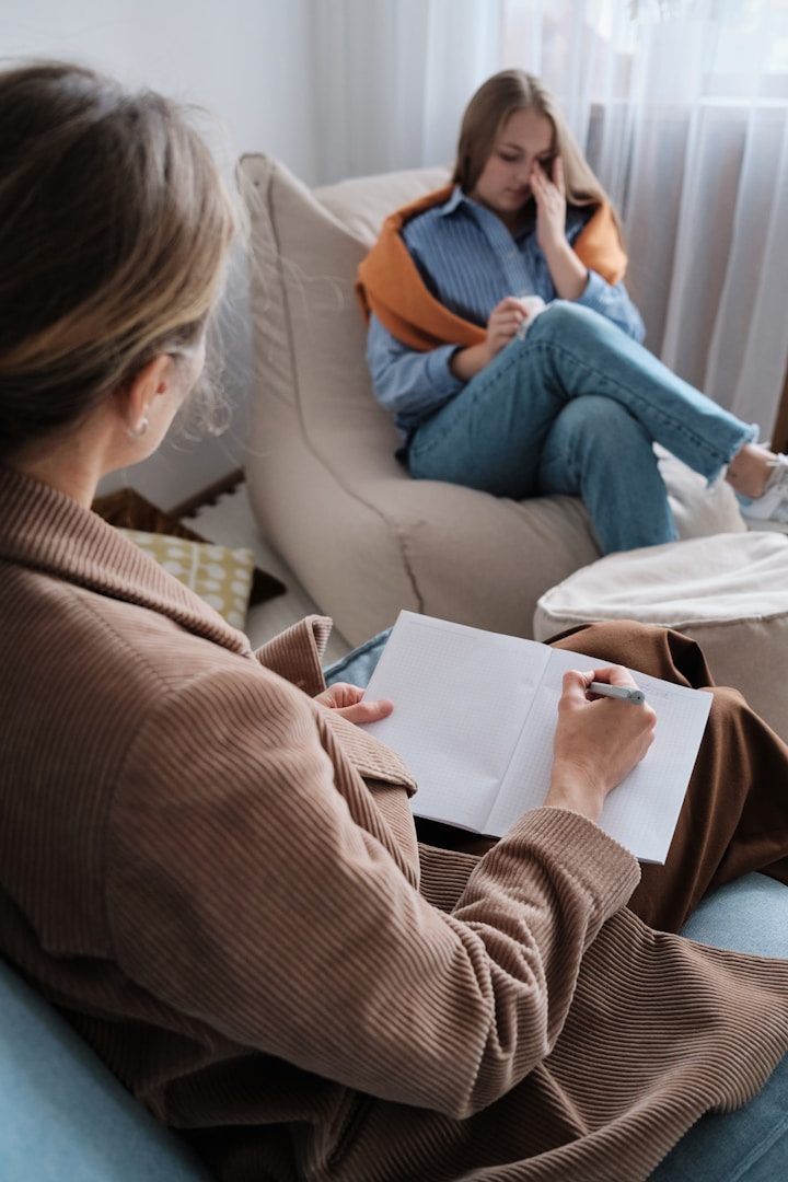 Therapie für Jugendliche Göppingen Eine Person sitzt auf einem Sessel und hält ein Notizbuch, während eine andere nachdenklich aussieht.